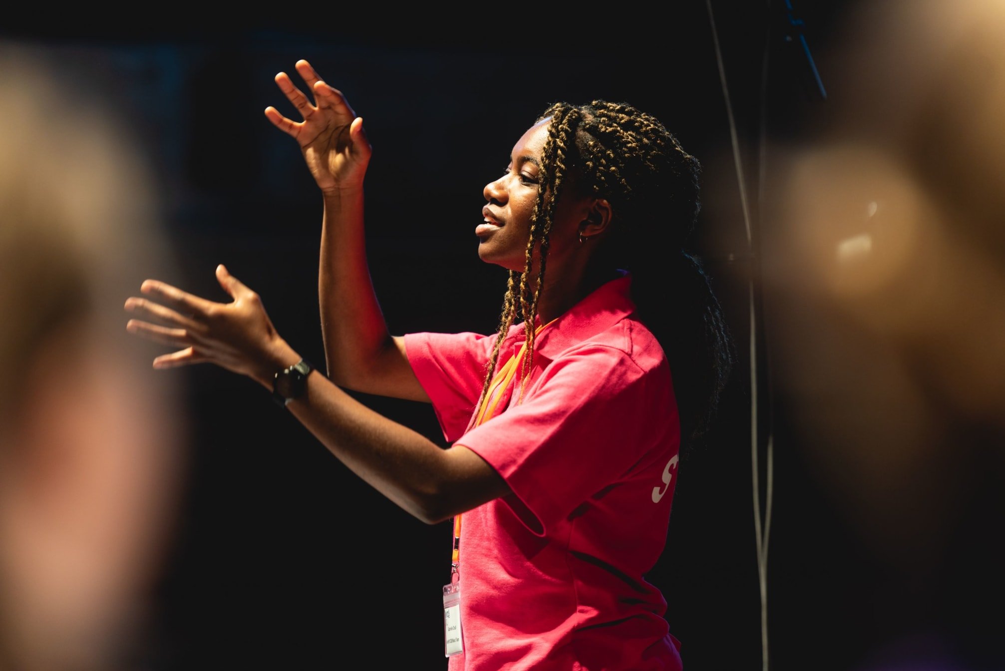 A black woman with long braids wearing a pink polo shirt conducts with her arms poised in midair