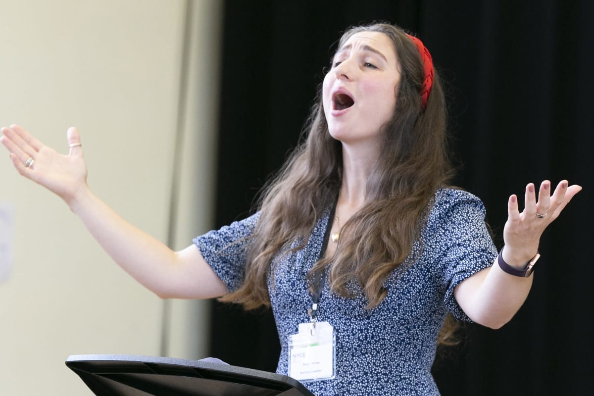 A white woman with long brown hair and a red hairband conducts with her arms outstretched