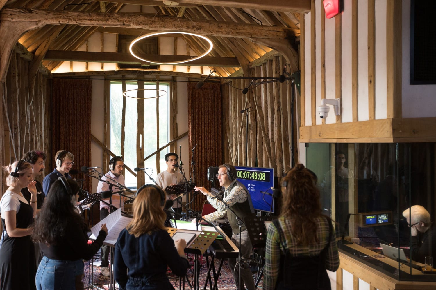Group of 8 singers and Ben Parry recording in a barn-style recording studio with exposed beams